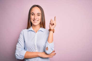 Zarif bir gömlek giyen genç ve güzel bir iş kadını pembe arka planda duruyor. Yüzünde kocaman bir gülümseme, eli ve parmağı kameraya bakıyor..
