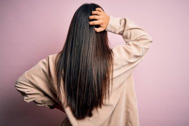 Genç, güzel Asyalı bir kadın pembe arka planda dikiliyor ve elleri kafasında şüpheyi düşünüyor.