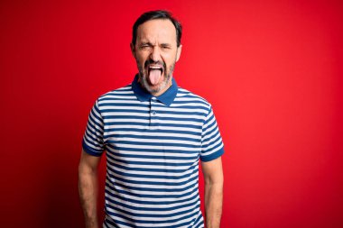 Orta yaşlı, beyaz, sıradan çizgili polo giyen, izole edilmiş kırmızı arka planda duran, komik bir ifadeyle dilini dışarı çıkaran bir adam. Duygu kavramı.