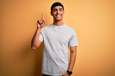 Yakışıklı Afro-Amerikan sporcu sarı arka planda spor giysisi giyiyor ve bir numarayı işaret ederken kendinden emin ve mutlu gülümsüyor..