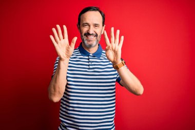 Orta yaşlı, beyaz, sıradan çizgili polo giyen, izole edilmiş kırmızı arka planda duran ve dokuz numarayı işaret eden, kendinden emin ve mutlu gülümseyen bir adam..