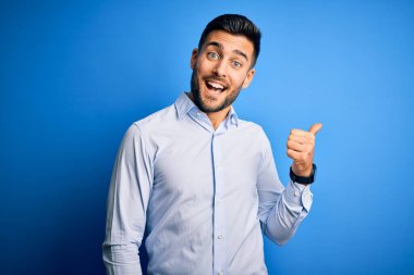 Zarif gömlekli genç adam mavi arka planda dikiliyor. Gülümsüyor, gülümsüyor ve başparmağıyla yana bakıyor..