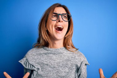 Genç, güzel, esmer bir kadın mavi arka plan üzerinde gözlük ve süveter takıyor deli deli deli bağırıyor ve bağırıyor agresif bir ifade ve kollar havada. Hayal kırıklığı konsepti.