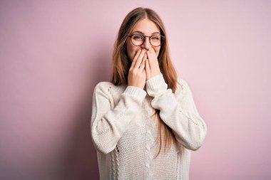 Genç, güzel, kızıl saçlı bir kadın pembe arka planda günlük süveter ve gözlük takıp gülüyor, kıkırdıyor, ağzı ellerle, dedikoduyla ve skandalla örtüyor.