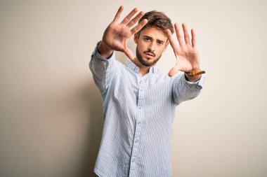 Sakallı, çizgili tişörtlü, beyaz arka planda duran, elleri ve parmakları kullanarak çerçeve yapan genç yakışıklı bir adam.