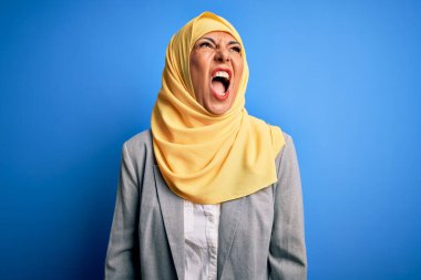 Orta yaşlı esmer iş kadını, geleneksel Müslüman tesettüre bürünmüş, mavi arka planda kızgın, kızgın ve öfkeli, öfkeyle bağıran bir kadın. Öfke ve agresif kavram.