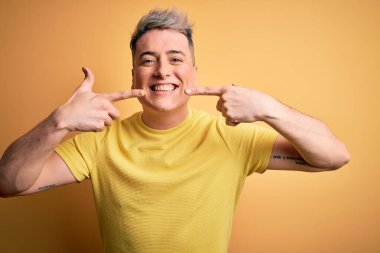 Sarı izole edilmiş arka planda sarı tişört giyen genç, yakışıklı, modern bir adam gülümseyen ve parmaklarıyla ve ağzıyla işaret eden. Diş sağlığı konsepti.