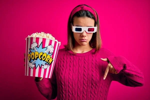 Young beautiful girl watching movie using 3d glasses eating box with popcorns Pointing down looking sad and upset, indicating direction with fingers, unhappy and depressed.