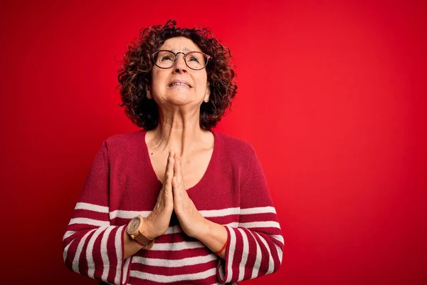 Middle age beautiful curly hair woman wearing casual striped sweater over red background begging and praying with hands together with hope expression on face very emotional and worried. Begging.