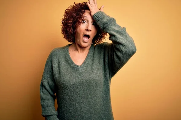 Middle age beautiful curly hair woman wearing casual sweater over isolated yellow background surprised with hand on head for mistake, remember error. Forgot, bad memory concept.