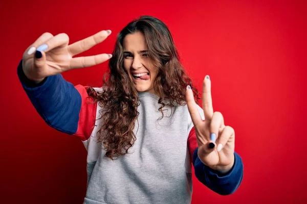 Young beautiful woman with curly hair wearing casual sweatshirt over isolated red background smiling with tongue out showing fingers of both hands doing victory sign. Number two.
