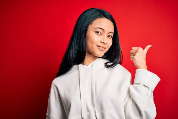 Young beautiful chinese sporty woman wearing sweatshirt over isolated red background smiling with happy face looking and pointing to the side with thumb up.