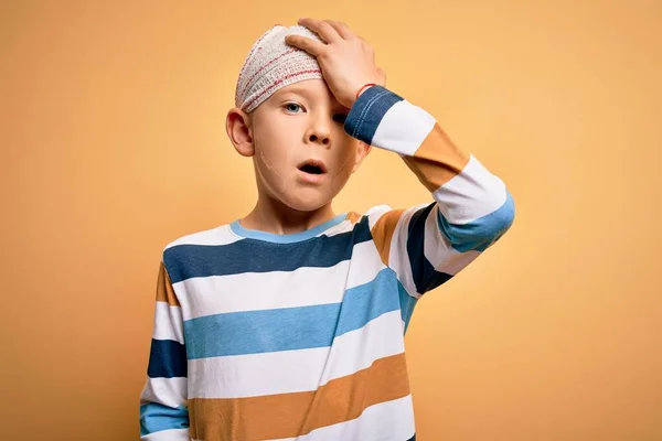 Young little caucasian kid injured wearing medical bandage on head over yellow background surprised with hand on head for mistake, remember error. Forgot, bad memory concept.