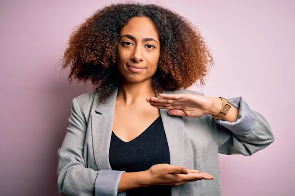 Young african american businesswoman with afro hair wearing elegant jacket gesturing with hands showing big and large size sign, measure symbol. Smiling looking at the camera. Measuring concept.