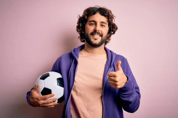 英俊的球员留着胡子踢足球拿着足球在粉红的背景下开心地笑着做个好手势 用手指竖起大拇指 很好的标志 — 图库照片