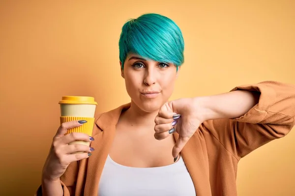 青いファッションの髪をした若い女性が怒った顔で黄色の隔離された背景にコーヒーを飲みます 親指で嫌いを示す負のサインダウン 拒絶概念 — ストック写真