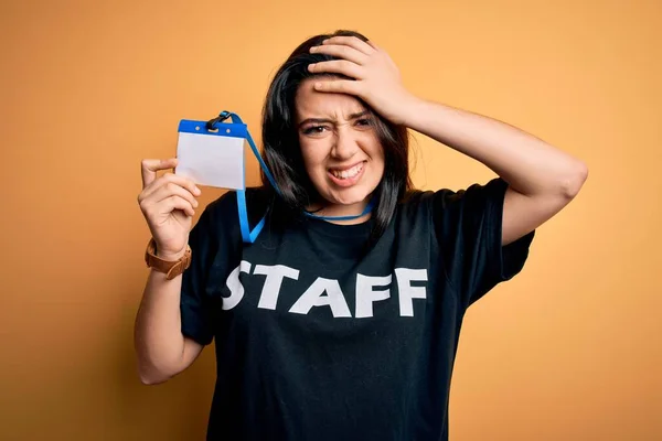 Young brunette worker woman wearing staff t-shirt as uniform showing id card stressed with hand on head, shocked with shame and surprise face, angry and frustrated. Fear and upset for mistake.