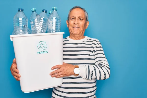 Senior man recycling holding trash can with plastic bottles to recycle over blue background with a happy face standing and smiling with a confident smile showing teeth