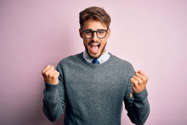 Young handsome man with beard wearing glasses and sweater standing over pink background celebrating surprised and amazed for success with arms raised and open eyes. Winner concept.