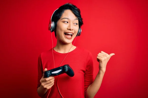 年轻美丽的亚裔游戏玩家女孩用操纵杆和耳机玩电子游戏 手指着 手指头着 笑容满面地出现在旁边 — 图库照片