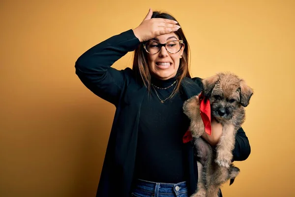 Young beautiful brunette woman holding cute puppy pet over isolated yellow background stressed with hand on head, shocked with shame and surprise face, angry and frustrated. Fear and upset for mistake.