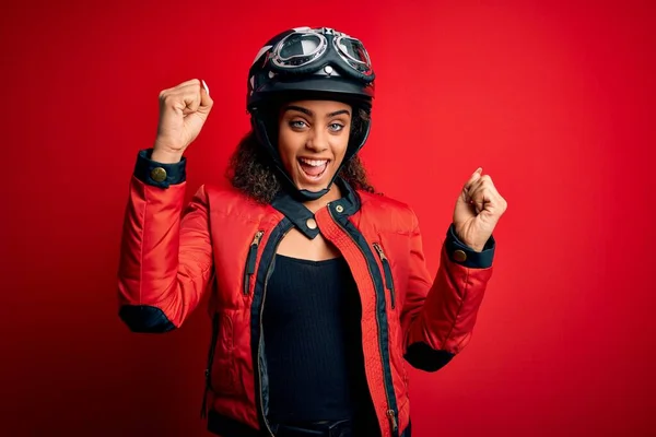 赤い背景の上に元ヘルメットと眼鏡をかけている若いアフリカ系アメリカ人のオートバイ愛好家の少女は 腕を上げて目を開けて成功したことに驚いて驚いています 勝者のコンセプト — ストック写真