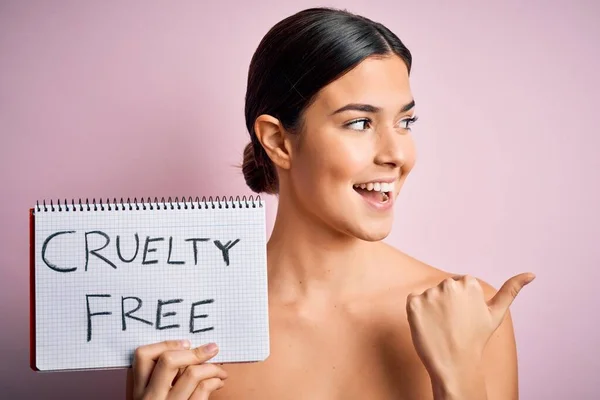 Young beautiful girl asking for cruelty free and vegan beauty cosmetics over pink background pointing and showing with thumb up to the side with happy face smiling