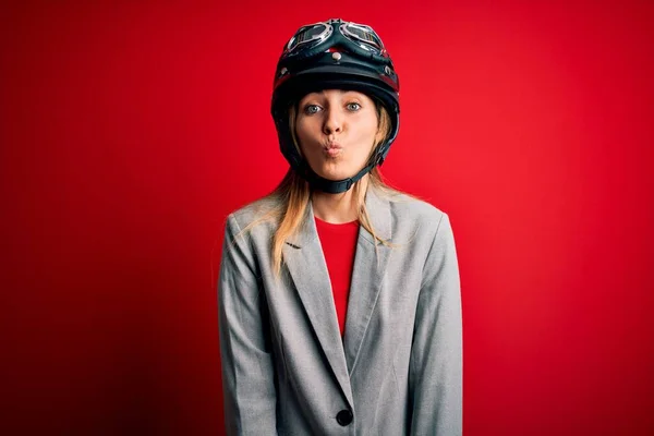若い美しいブロンドのオートバイの女性は 狂気とコミカルなジェスチャーで魚の顔を作る赤い背景の上にオートバイのヘルメットを着ています 面白い表現 — ストック写真