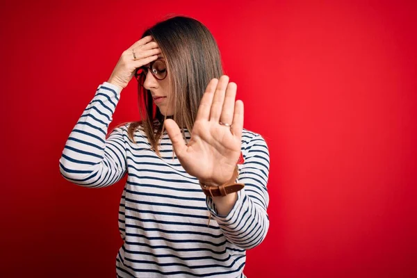 Young beautiful blonde woman with blue eyes wearing glasses standing over red background covering eyes with hands and doing stop gesture with sad and fear expression. Embarrassed and negative concept.