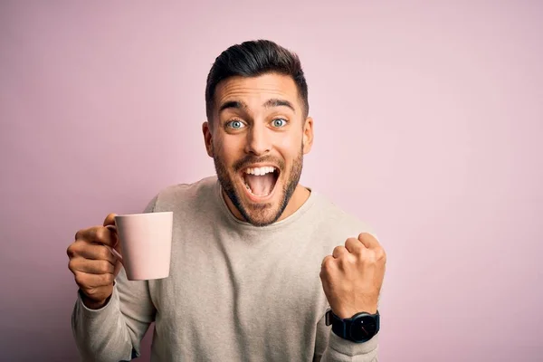 Young handsome man drinking a cup of hot coffee over pink isolated background screaming proud and celebrating victory and success very excited, cheering emotion