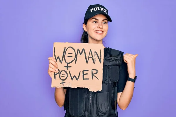 Police woman wearing security bulletproof vest uniform holding woman power protest cardboard pointing and showing with thumb up to the side with happy face smiling