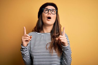 Genç, güzel esmer kadın Fransız beresi takıyor ve sarı arka planda gözlük takıyor şaşkın şaşkın yukarı bakıyor ve parmaklarıyla işaret ediyor ve kollarını kaldırıyor..