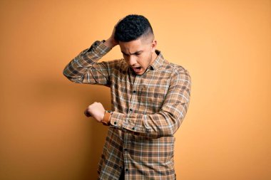 Sarı arka planda duran sıradan tişörtlü genç yakışıklı adam saatin saatine bakıyor endişeli, geç kalmaktan korkuyor.