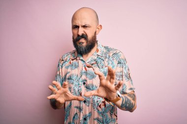 Sakallı ve dövmeli yakışıklı bir adam pembe arka planda sıradan bir çiçek gömleği giyiyor, tiksindirici ve tiksindirici bir yüz ifadesi takınıyor..