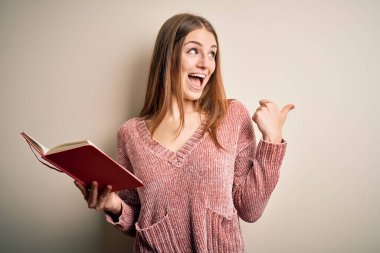 Genç, güzel, kızıl saçlı, kitap okuyan, izole edilmiş beyaz arkaplanı olan, gülümseyen yüzüyle baş parmağıyla gösteren bir kız öğrenci.