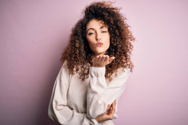 Kıvırcık saçlı ve piercingli güzel bir kadın pembe arka planda sıradan bir süveter giyiyor. Kameraya bakıyor ve havada elle öpüşüyor. Güzel ve seksi. Aşk ifadesi.