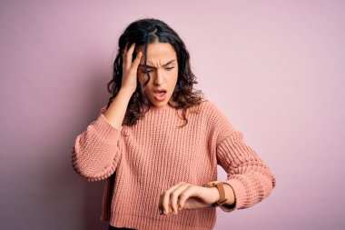 Kıvırcık saçlı, günlük süveter giyen, soyutlanmış pembe arka planda olan genç ve güzel bir kadın. Endişeli saate bakıyor, geç kalmaktan korkuyor.