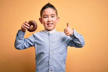 Sağlıksız çikolatalı donut yiyen küçük çocuk izole edilmiş sarı arka planda mutlu bir şekilde gülümsüyor.
