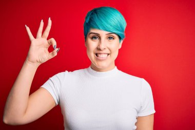 Mavi saçlı, kırmızı arka planda sıradan tişörtler giyen güzel bir kadın gülümseyerek el ve parmaklarıyla tamam işareti yapıyor. Başarılı ifade.