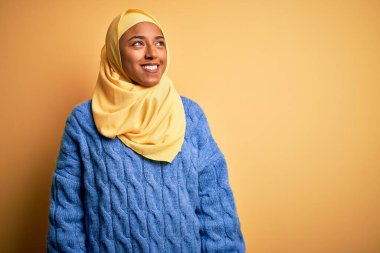 Genç Afrikalı Afro kadın, izole edilmiş sarı arka plan üzerinde Müslüman başörtüsü takıyor. Yüzünde gülümseme, doğal bir ifade ile yan yana bakıyor. Kendinden emin gülen.