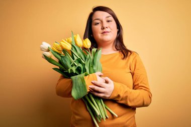 Sarı arka planda romantik bir buket lale tutan güzel ve cüsseli bir kadın yüzünde ciddi bir ifadeyle rahatlamış. Basit ve doğal bir şekilde kameraya bakıyorsun..