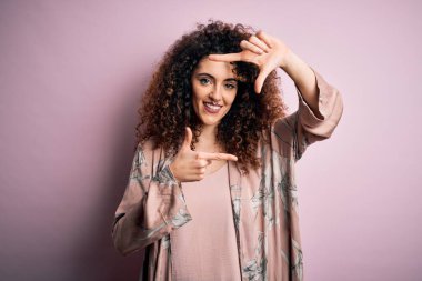 Kıvırcık saçlı, piercingli, sıradan pembe tişörtlü, gülümseyen, elleri ve parmakları mutlu yüzlü güzel bir kadın. Yaratıcılık ve fotoğrafçılık kavramı.