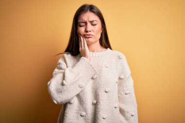 Mavi gözlü genç bir kadın sarı arka planda dikilmiş diş ağrısı ya da diş hastalığından dolayı acı dolu bir ifadeyle ağıza dokunuyor. Dişçi