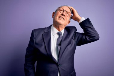 Gri saçlı, gözlüklü, şık takım elbiseli ve mor arka planda kravat takan, kendine güvenen, el hareketleriyle saçlarına dokunan, çekici ve modaya uygun poz veren kıdemli iş adamı.