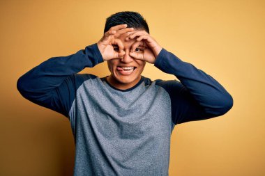 Sarı arka plan üzerinde sıradan tişörtler giyen genç, yakışıklı latin adam dürbün gibi jest yapıyor, gözleri parmaklarından bakıyor. Çılgın ifade.