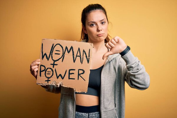 Young beautiful sportswoman holding woman power banner over isolated yellow background with angry face, negative sign showing dislike with thumbs down, rejection concept