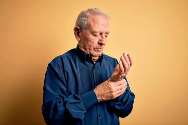 Grey haired senior man wearing casual blue shirt standing over yellow background Suffering pain on hands and fingers, arthritis inflammation