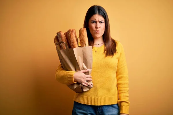 年轻美丽的女人拿着一袋新鲜健康的面包 披着黄色背景的怀疑和紧张 因为问题皱着眉头 消极的人 — 图库照片