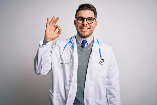 青い目をした若い医者の男は孤立した背景の上に医療用のコートと聴診器を身に着けていて 手と指でOkサインをします 成功した表現 — ストック写真