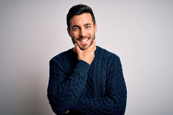 Young handsome man with beard wearing casual sweater standing over white background looking confident at the camera smiling with crossed arms and hand raised on chin. Thinking positive.